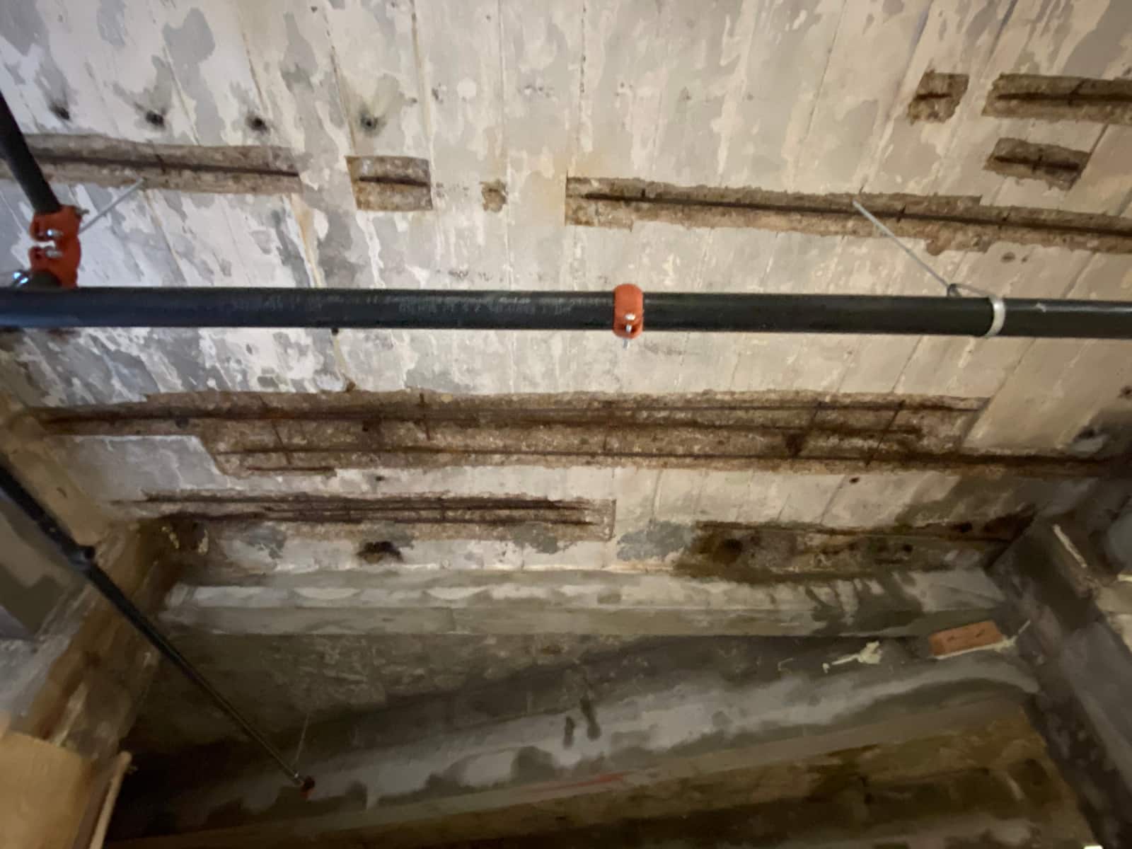 Slab soffit with long chipped strips exposing reinforcing bars and a pipe below.