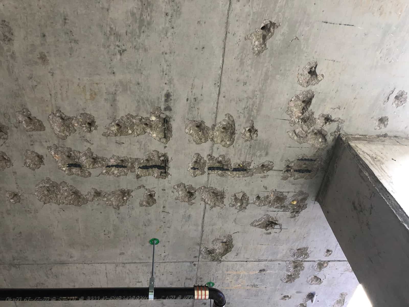 Chipped slots in a slab soffit exposing dark embedded bar segments near a column.