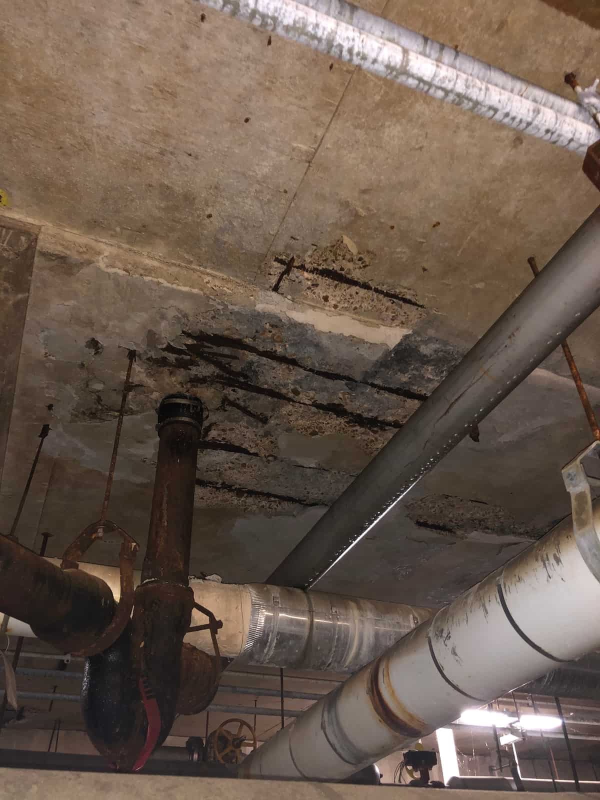 Slab underside with a spalled area and exposed rebar above piping.