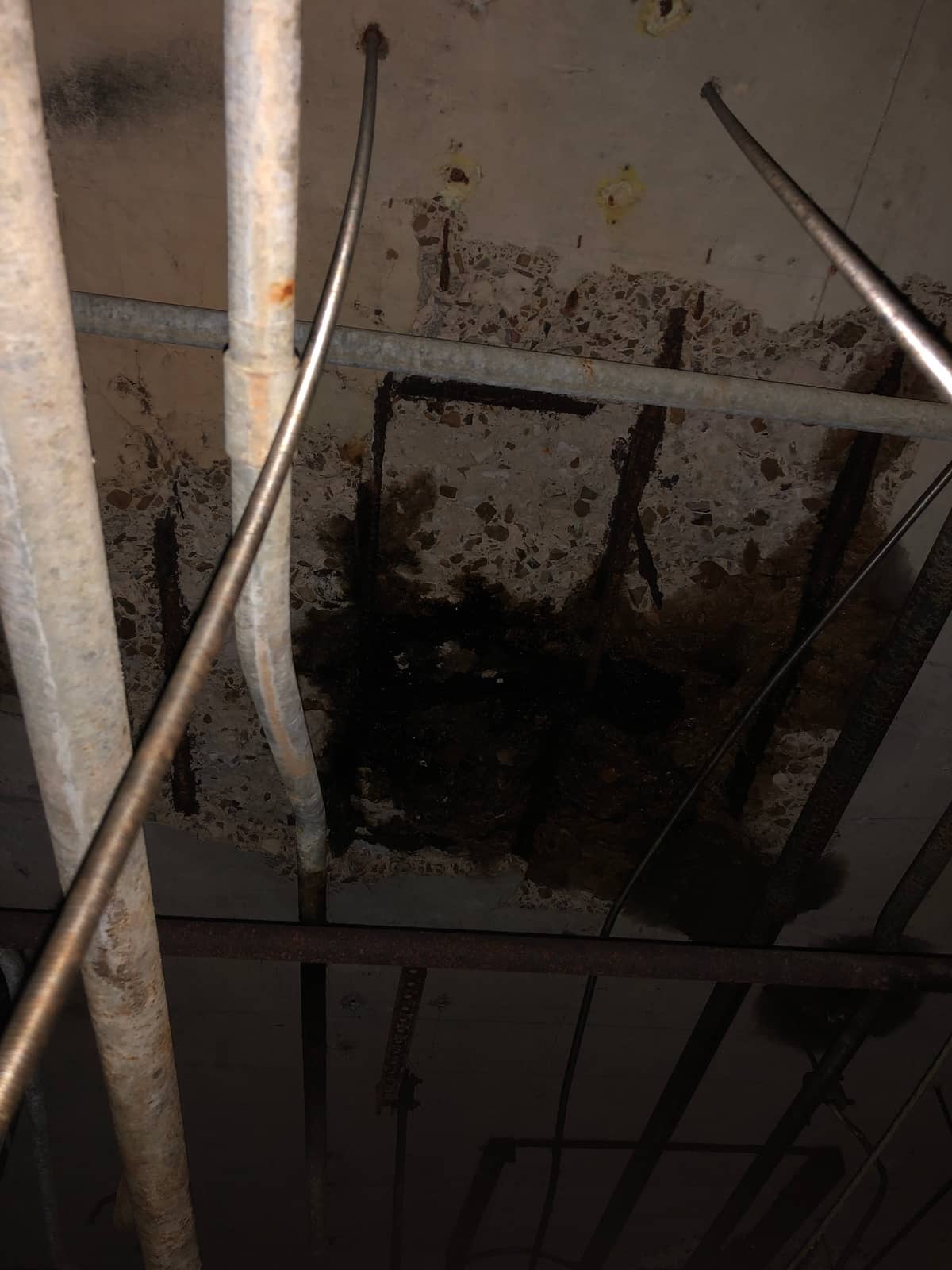 Slab underside with heavy spalling and exposed corroded rebar above pipes.