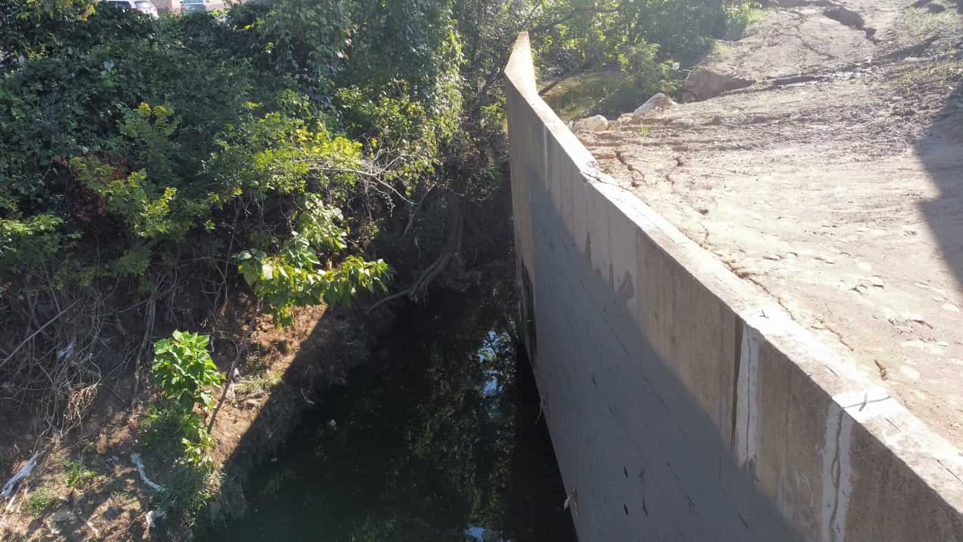 Concrete channel retaining wall with cracks above water and vegetation.