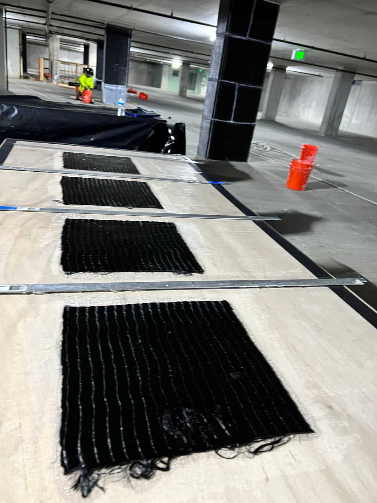 Saturated carbon fiber sheets laid out on a work table in a parking garage, wrapped columns behind.