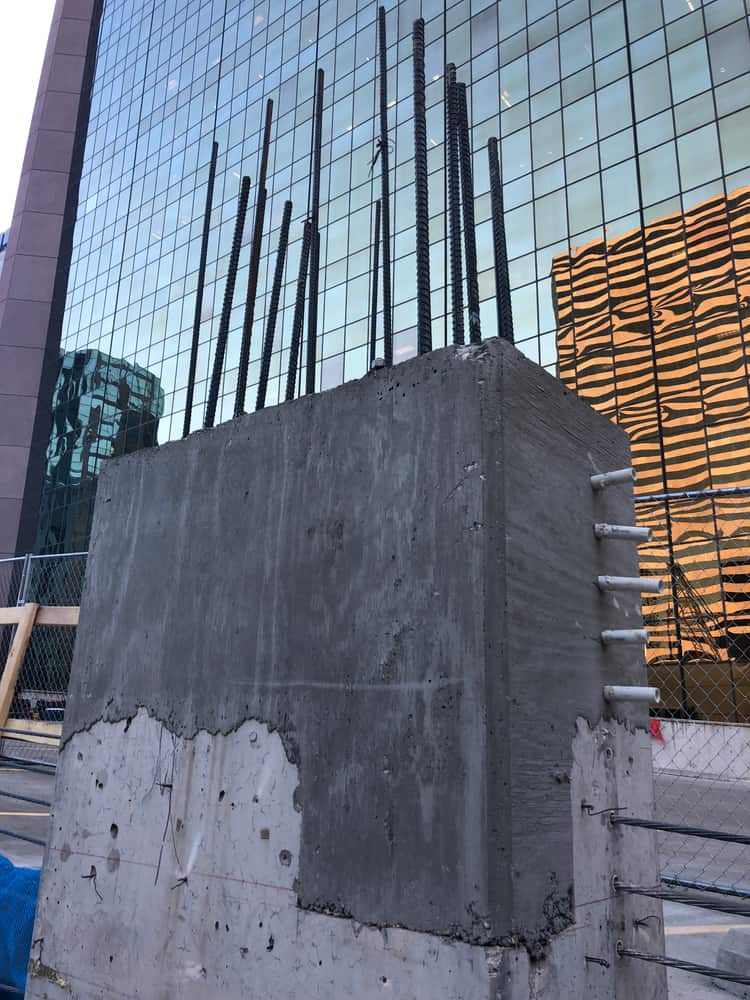 New concrete column extension with protruding rebar against a glass tower.