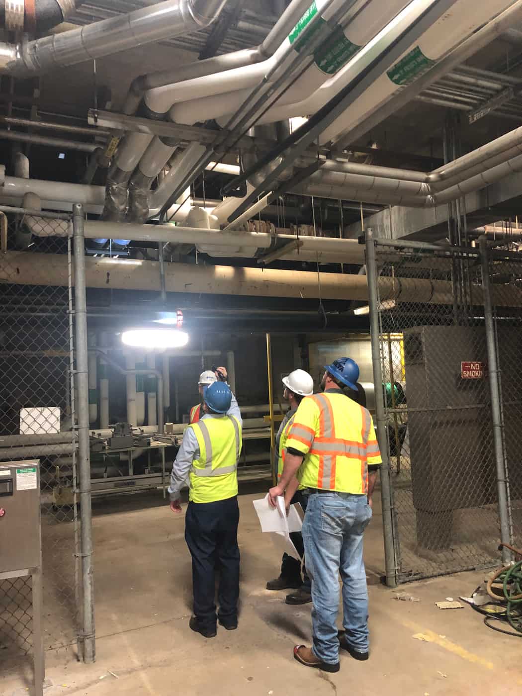 Inspectors in hard hats and vests looking up at piping in a mechanical room.