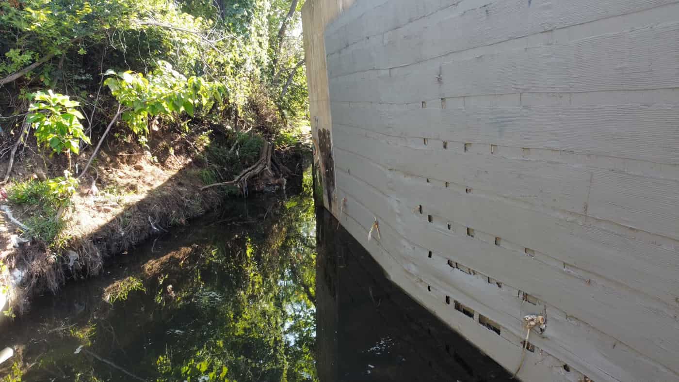 Board-formed concrete wall rising from creek water beside vegetation.