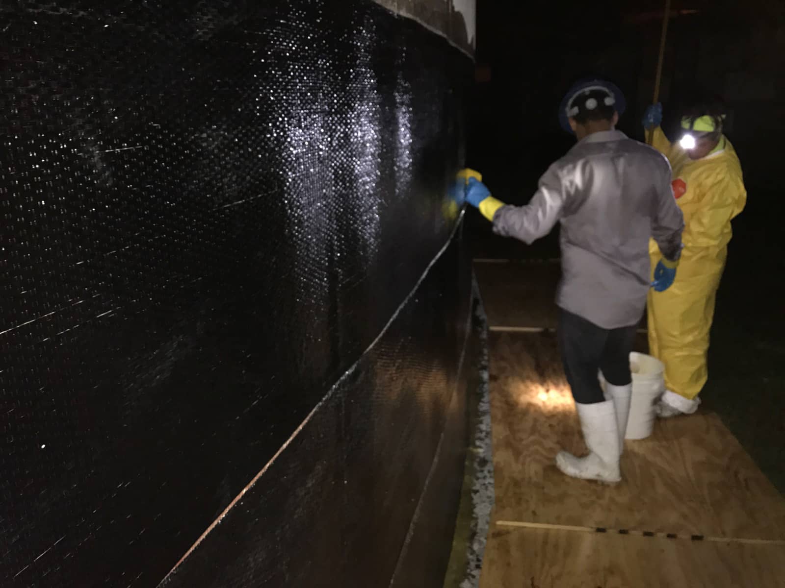 Night work applying black carbon fiber fabric to a curved concrete tank wall.