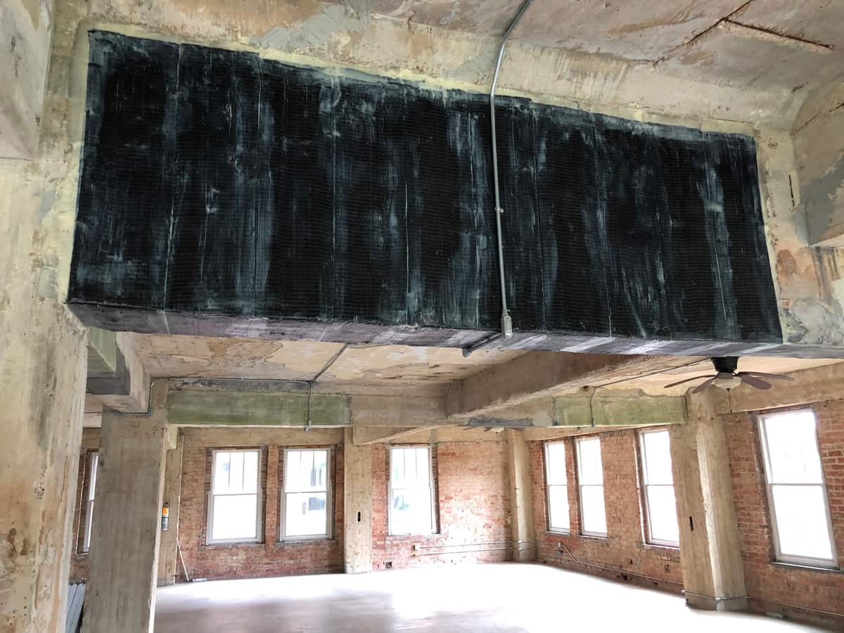 Deep concrete beam wrapped in black saturated carbon fiber fabric above an empty room.