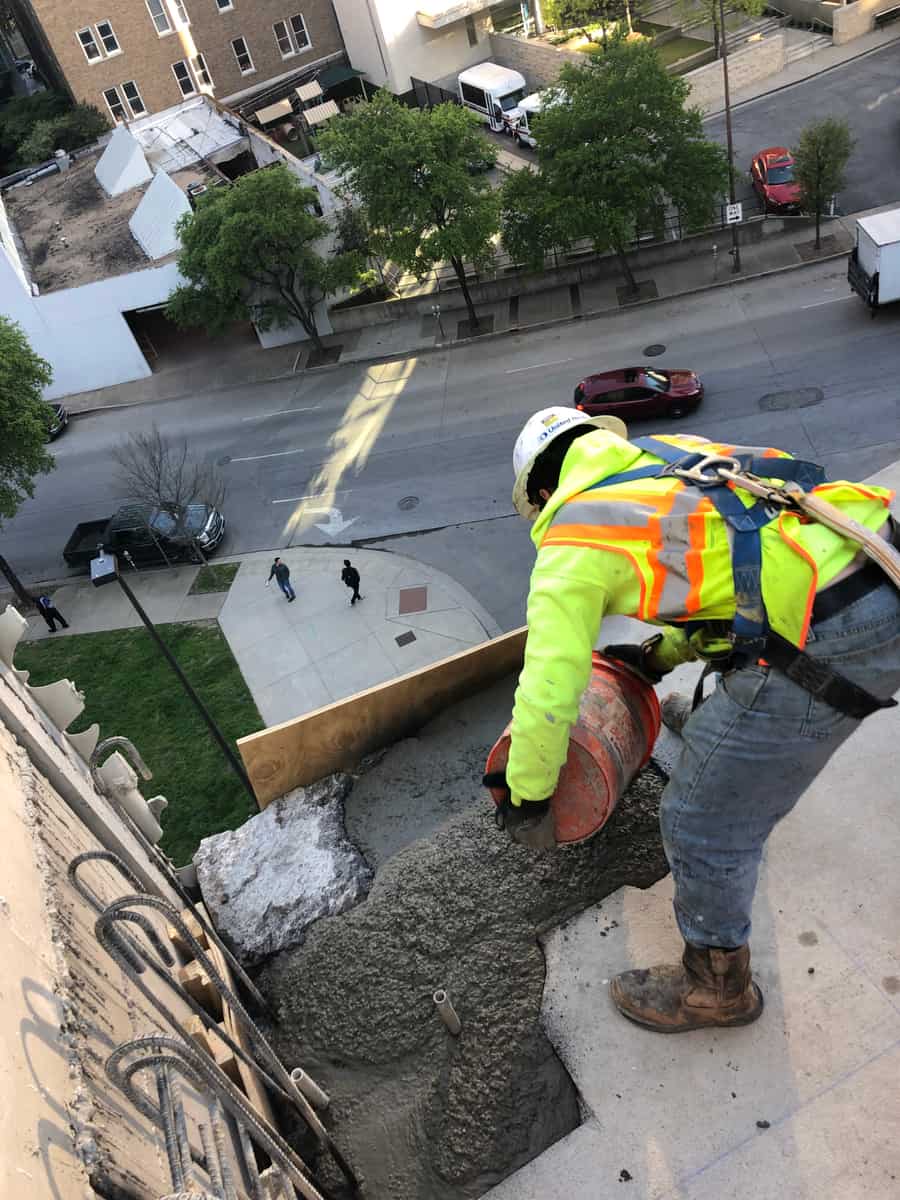 Harnessed worker placing concrete at a high ledge overlooking a street.