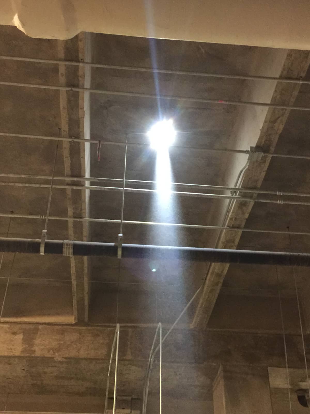 Underside of raked stadium seating beams with pipes and a work light.