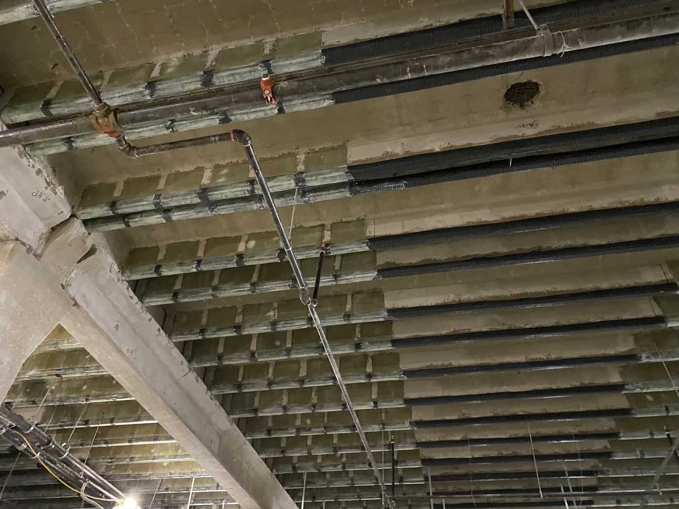 Angled view of pan-joist slab underside with dark carbon fiber strips and a sprinkler pipe.