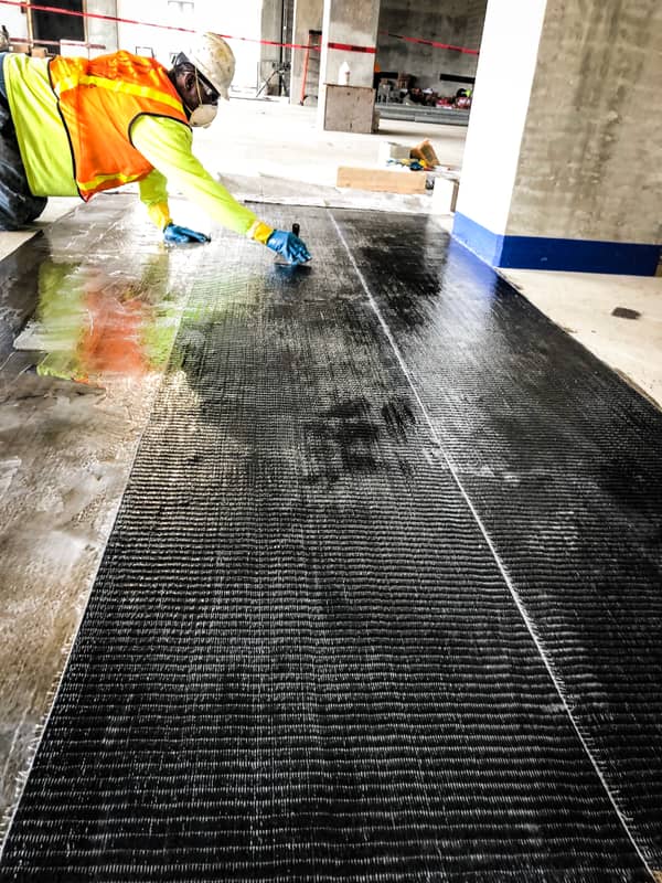 Worker rolling a carbon fiber sheet onto a concrete deck near a column.