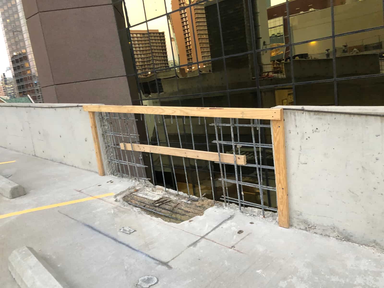Parapet opening with an exposed rebar mat and a timber rail on a parking deck.