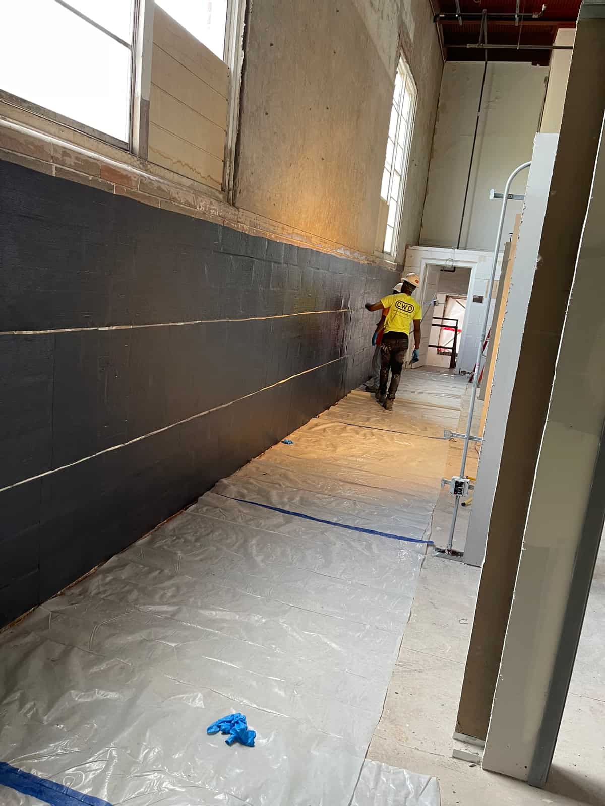 Black composite applied along a tall masonry wall with plastic sheeting on the floor.