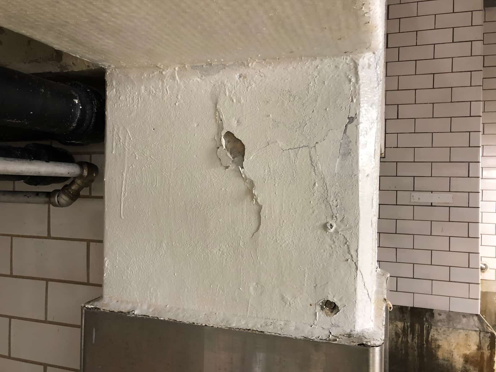 White painted column with cracks and small spalls next to a tile wall.