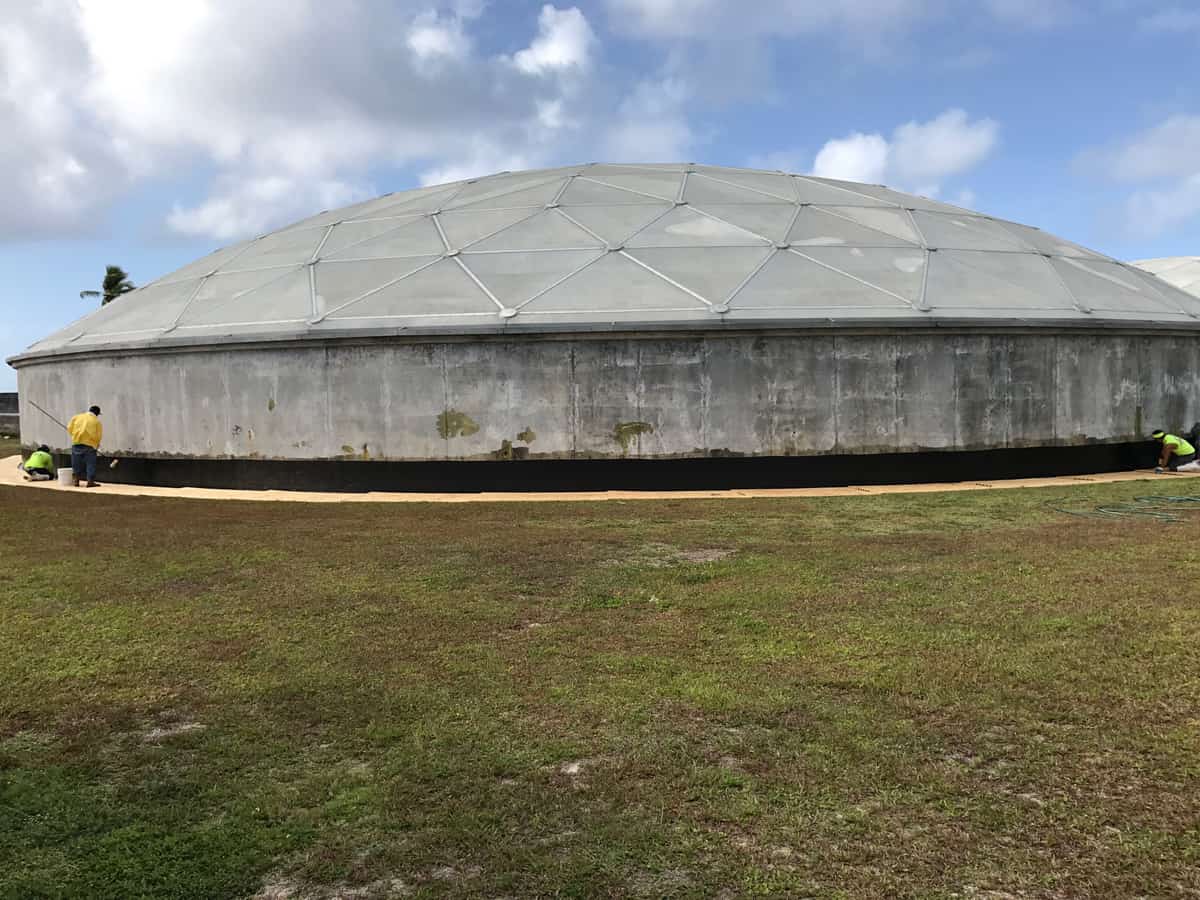 Domed concrete tank with workers applying a dark composite band around the base of the wall.