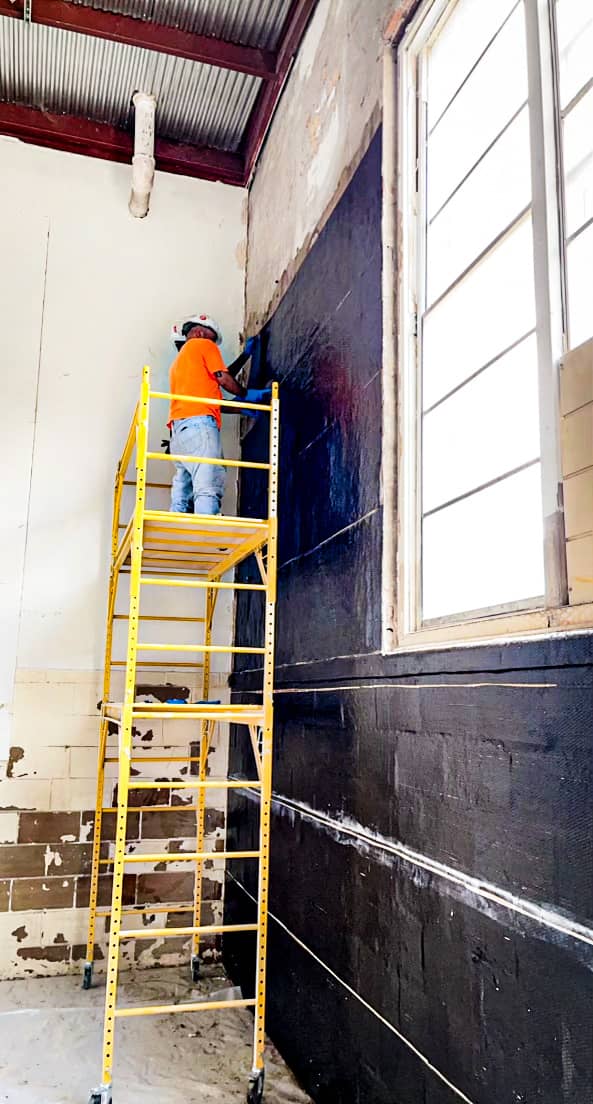 Worker on a yellow scaffold applying black composite to a tall masonry wall beside windows.